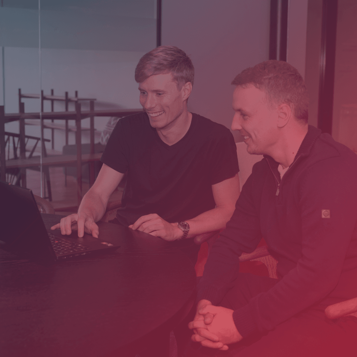 Two men smiling and looking at a laptop screen while sitting at a round table in a modern office setting.