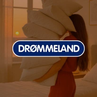 En kvinde bærer på flere puder i soveværelset med solnedgang udenfor, med et "drømmeland" logo tydeligt placeret i forgrunden af billedet